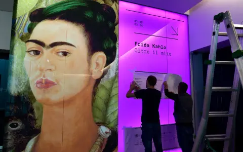 Un portrait de Frida Kahlo, avec deux hommes installant une affiche pour une exposition sur son histoire.