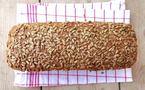Ein rustikales Brot, bedeckt mit vielen Sonnenblumenkernen, liegt auf einem karierten Küchentuch.