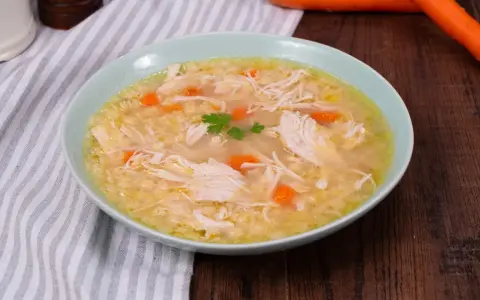 Un confortevole brodo di pollo con pezzi di pollo sfilacciato, carote e un rametto di prezzemolo, servito in una ciotola azzurra.