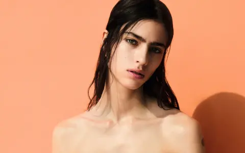 A striking portrait of Loli Bahia, her dark hair cascading around her shoulders against a vibrant peach backdrop.