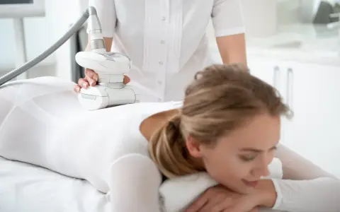 Kobieta w białym stroju poddaje się zabiegowi endermologii LPG. Widoczne są ręce terapeuty trzymające głowicę urządzenia, które masuje nogę pacjentki. Efekty zabiegu są obiecujące.