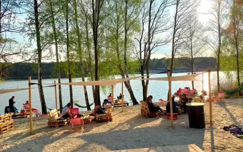 Pola namiotowe nad jeziorem, ludzie odpoczywają na plaży pod baldachimami, wśród drzew.