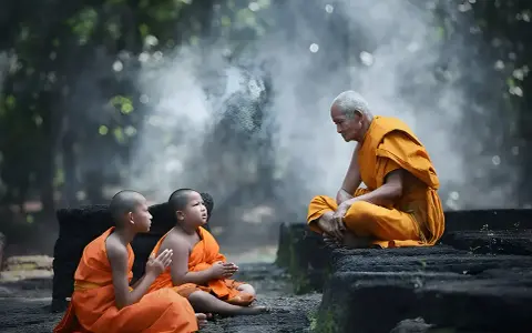 Äldre munk i orange kläder undervisar två unga munkar i en lugn miljö, en scen av buddhistisk puja.