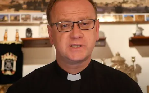Wojciech Węgrzyniak w okularach, w czarnej sutannie z białą kolaretką, przemawia. W tle widać zwoje Tory i obrazy.
