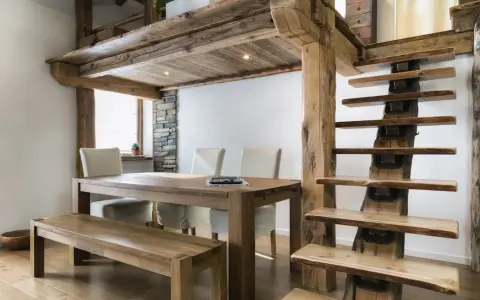 Une salle à manger rustique avec une table en bois, des chaises et un banc, sous une mezzanine maison ancienne aux poutres apparentes. L'escalier en bois brut complète le décor.