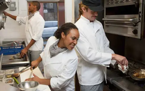 La brigada de cocina y sus funciones: chefs preparan alimentos, uno corta vegetales, otro cocina en sartén y otro lava utensilios.