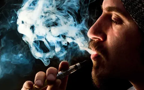 Hombre vapeando, el vaper sin nicotina es malo. El humo azul forma figuras fantasmagóricas.