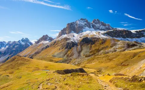 Ampia **foto grandangolo** di montagne innevate e pendii dorati autunnali, con un sentiero che serpeggia tra l'erba.