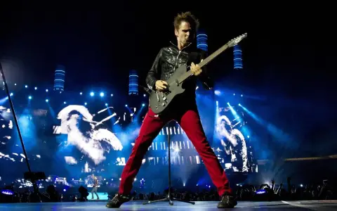 Matt Bellamy de Muse España, con pantalones rojos y chaqueta negra, toca la guitarra en un escenario iluminado por luces azules y pantallas gigantes.