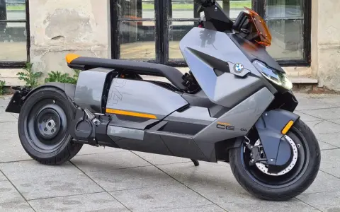 Szary skuter elektryczny BMW CE 04 z pomarańczowymi akcentami stoi na bruku przed budynkiem z dużymi oknami.