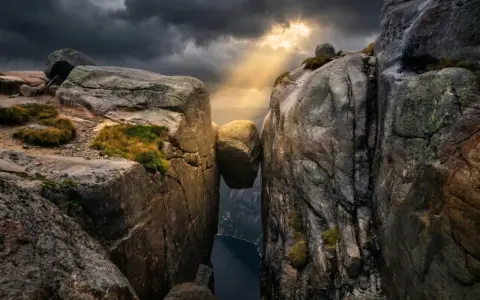 Enorm sten balanserar mellan klippor i Norge. Dramatiskt ljus bryter igenom molnen över en djup fjord.