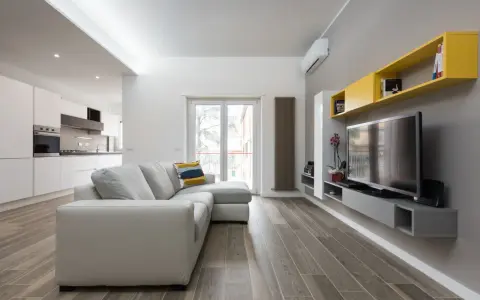 Un progetto soggiorno stretto e lungo con divano grigio, TV a parete e mensole gialle. La cucina moderna è visibile sullo sfondo.