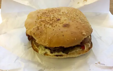 Burger z sezamem na białym papierze, widoczne składniki: mięso, sos, warzywa.