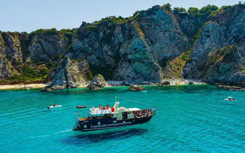 Barca turistica naviga nelle acque cristalline vicino al porto di Tropea, con imponenti scogliere a picco sul mare e piccole spiagge affollate.