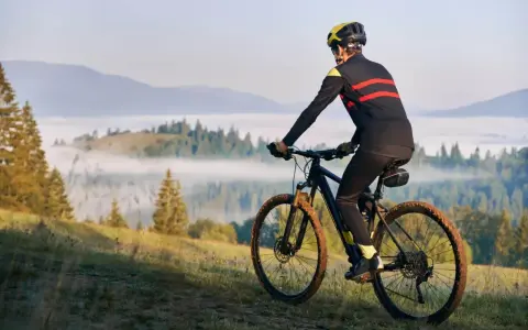 Rowerzysta w kasku i sportowym stroju pokonuje górski szlak, utrzymując dynamiczną kadencję rower. Mgła spowija doliny, tworząc malowniczy krajobraz.