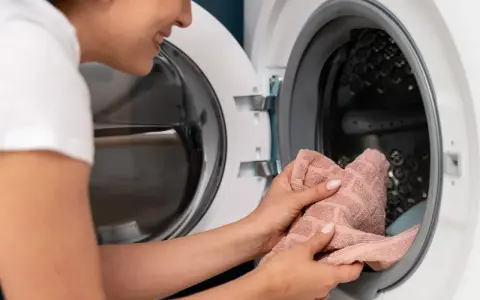 Uśmiechnięta kobieta wkłada różowy ręcznik do pralki. Zastanawia się, ile trwa pranie w 60 stopniach.