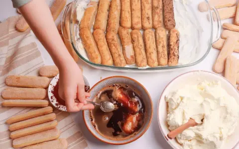 Dziecko zanurza biszkopty w kawie, przygotowując warstwy do tiramisu. Obok krem i kakao.