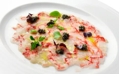 Delicado carpaccio de gambas con caviar negro, aceite de oliva y brotes verdes, presentado en un plato blanco.