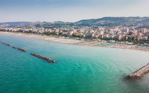 Vista aerea di San Benedetto del Tronto: una lunga spiaggia con ombrelloni colorati e il mare turchese, con dighe frangiflutti che proteggono la costa.