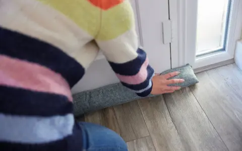 Ocieplanie drzwi wejściowych: osoba w kolorowym swetrze układa podkładkę izolacyjną pod drzwiami, by zatrzymać przeciągi.