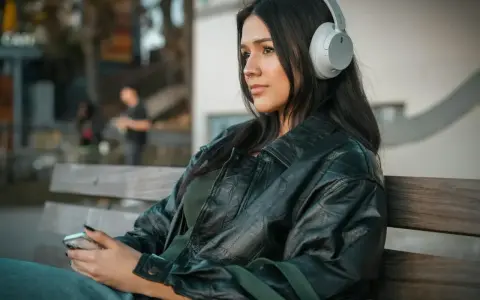 Młoda kobieta w słuchawkach, słuchająca podcastu o wymowie francuskiej, siedzi na ławce, trzymając telefon.