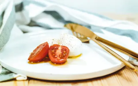Kuleczki mozzarelli z pomidorkami koktajlowymi na białym talerzu, obok złote sztućce i ściereczka.