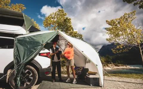 Zwei Frauen genießen das Frauen Camping Abenteuer am See. Ein Zelt ist am Auto befestigt, mit Blick auf Berge und Wasser.