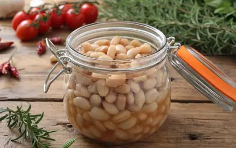Ein Glas mit dicken Bohnen, bereit zum Einkochen, steht auf einem Holztisch. Daneben liegen Rosmarin, Tomaten und getrocknete Chilis.