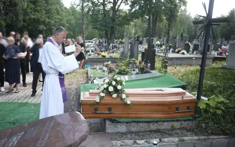 Wojciech Magnuski żegna zmarłego na cmentarzu. Ksiądz w białej szacie czyta z księgi przy trumnie ozdobionej białymi różami.