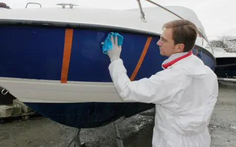 Un homme en combinaison blanche effectue l'entretien d'un bateau bleu et blanc, le lustrant avec un chiffon bleu.