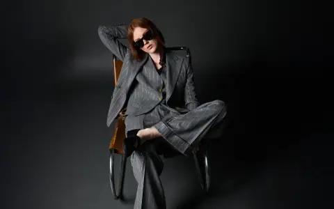 Donna con outfit giacca doppiopetto donna a righe grigie, occhiali da sole, seduta su una poltrona di pelle.