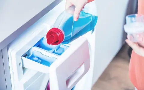 Eine Hand gießt flüssiges Waschmittel in das Fach einer Waschmaschine. So benutzt man eine Waschmaschine richtig.