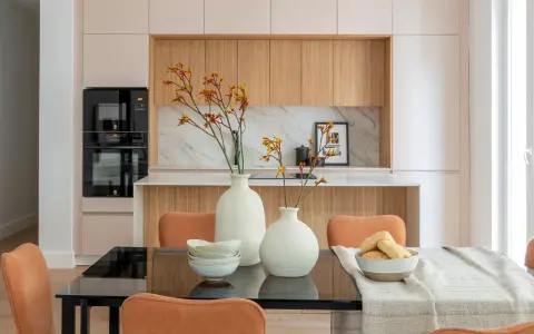 Dos jarrones blancos con flores anaranjadas y amarillas, cuencos apilados y un bol con panecillos son elementos decorativos que poner encima de una mesa de comedor.