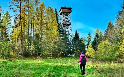 Spacerowiczka w różowej kurtce idzie przez łąkę w kierunku wieży widokowej. W tle jesienne lasy i szczyty górskie Muszyny.