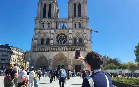 Una persona toma una foto de la fachada de Notre Dame. Mucha gente pasea por la plaza, disfrutando de su visita a Notre Dame.