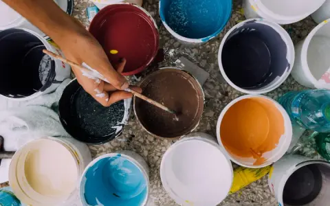 Ręka z pędzlem miesza farby w wiaderkach. Widać różne kolory, ale też potencjalne wady farby ceramicznej, jak nierównomierne rozprowadzenie.