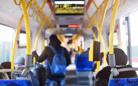 Wnętrze autobusu z żółtymi poręczami. Młody mężczyzna z niebieskim plecakiem idzie do tyłu. Uważaj, by nie dostać mandatu za brak biletu.