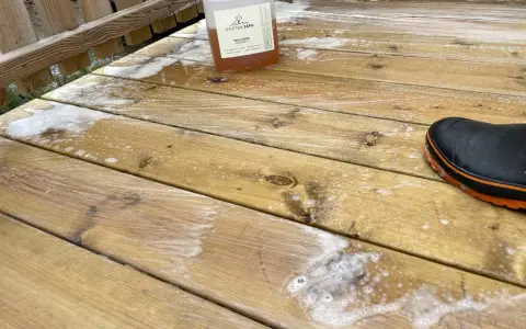 En flaska linoljesåpa står på en altan som håller på att rengöras. En svart stövel med orange kant syns i nedre högra hörnet.