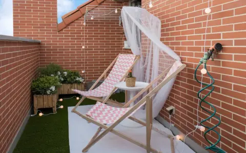 Acogedora terraza de 10 metros cuadrados con dos tumbonas, plantas y luces decorativas. Ideal para relajarse.