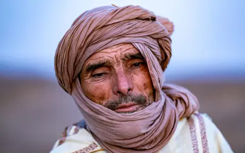Porträt eines alten Berber Hirtennomaden mit einem Turban und traditioneller Kleidung, dessen Gesicht von der Sonne gezeichnet ist.