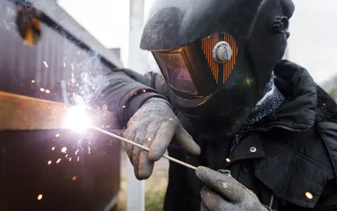 Soldador con casco y guantes usa un electrodo para unir metales, creando chispas brillantes y humo.