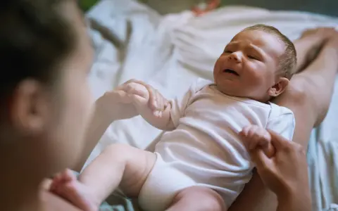 Płaczliwe niemowlę w białym body, trzymające mamę za rękę. Co robić, gdy pojawia się histeria u niemowlaka?