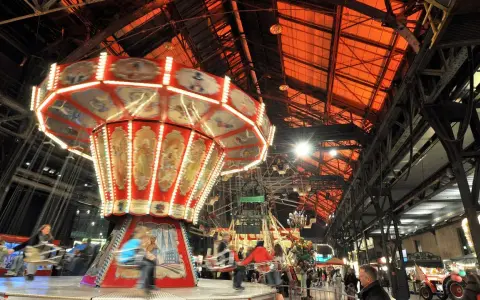Drehende Kettenkarussells und bunte Lichter auf einem historischen Jahrmarkt in einer alten Industriehalle.