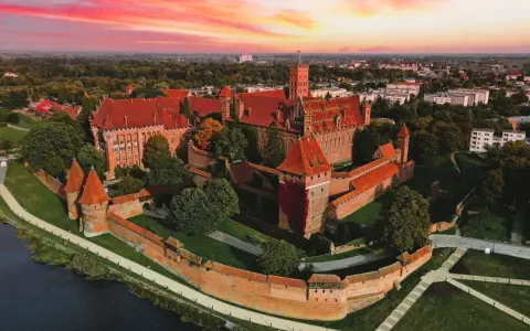 Zamek krzyżacki w Malborku, imponująca twierdza z czerwonej cegły, otoczona fosą i zielenią, pod rozgwieżdżonym, różowym niebem. Ciekawostki o jego historii czekają na odkrycie.