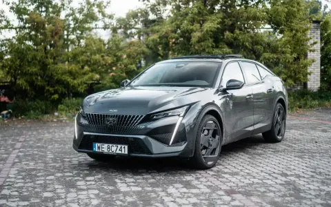 Nowoczesny Peugeot 408 Hybrid w ciemnoszarym kolorze, zaparkowany na brukowanym podjeździe, otoczony zielenią.
