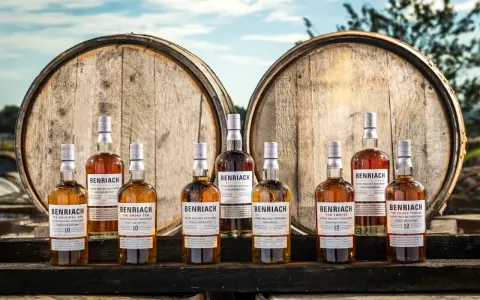 Butelki whisky BenRiach stoją przed beczkami, które zdradzają, skąd pochodzi whisky.