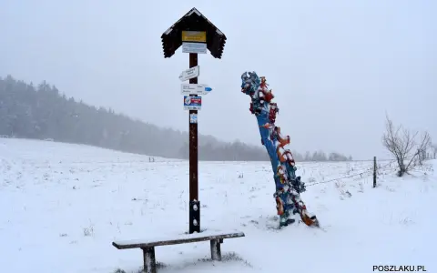 Zimowy krajobraz z ławką i drogowskazem turystycznym. Obok kolorowa rzeźba, być może symbolizująca skopiec szczyt.