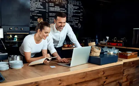 Pareja revisa menú en laptop, planificando su negocio de hostelería en España.