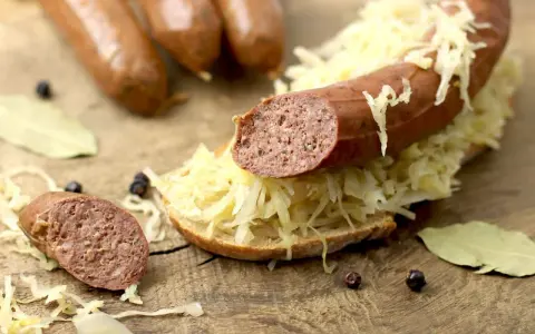 Lungenwurst zubereiten: Eine Scheibe Brot mit Sauerkraut und Lungenwurst belegt, daneben weitere Würste, Gewürze und Lorbeerblätter auf Holzuntergrund.