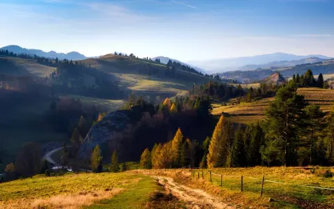 Malowniczy krajobraz górski z krętą drogą, drzewami i łagodnymi wzgórzami w jesiennym słońcu.